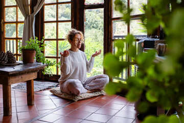 Full length attentive young Caucasian woman making mudra gesture, sitting in lotus position sitting on soft cushion on floor at home. Peaceful woman meditating deeply, doing breathing yoga 