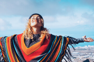 Blonde woman in hat and colorful poncho relaxing with open arms in blue winter sky. People's freedom style. © simona