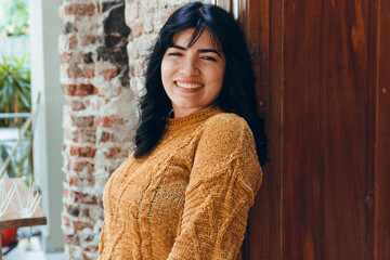 waist up portrait, young pretty latin woman smiling happy watching camera posing next to wooden door