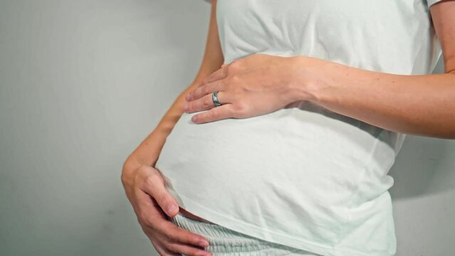 A young pregnant girl with a large stomach, stands at home against a white wall and touches her stomach with her hands.