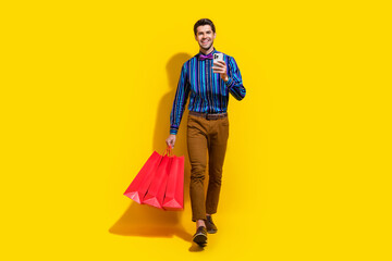 Full size photo of clever guy dressed striped shirt go with shopping bags order clothes on smartphone isolated on yellow color background