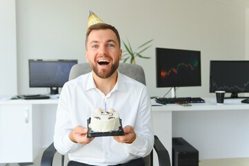 Young happy businessman celebrating his birthday in the office. He holds a cake in his hands.