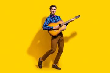 Full size photo of optimistic funky guy dressed striped shirt playing on acoustic guitar on stage isolated on yellow color background