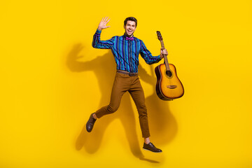 Fototapeta na wymiar Full size photo of pleasant guy dressed striped shirt jumping hold guitar waving palm say hello isolated on yellow color background