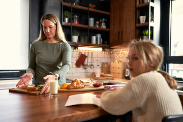 A little girl, pencil in hand, works on homework as her mother assists, blending breakfast and learning into a delightful morning routine.