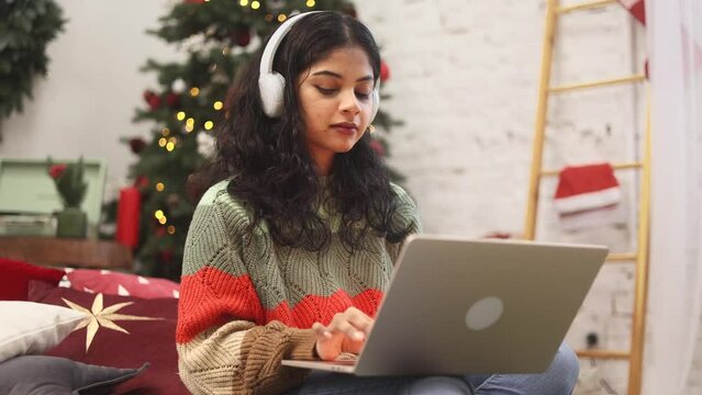 Charming indian woman relaxing at home listening music or working online browsing products in internet store reading news or social media profile on laptop computer near Christmas tree indoors