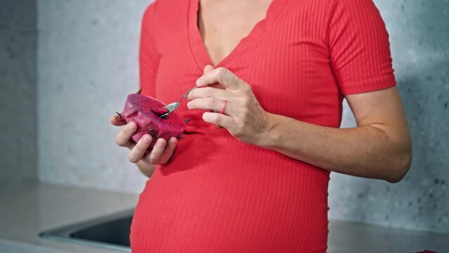 A young pregnant girl with a large stomach, stands at home in a colorful dress and eats dragonfruit.