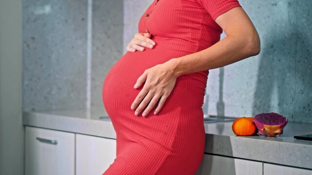 A young pregnant girl with a large stomach, stands in the kitchen in a colorful dress and touches her stomach with her hands.