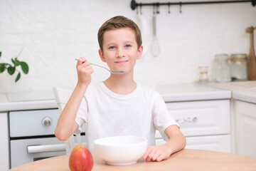 Little boy eating cereal with milk, apple for breakfast at wooden table in light white kitchen.