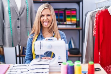 Naklejka na ściany i meble Blonde woman dressmaker designer using sew machine sticking tongue out happy with funny expression. emotion concept.