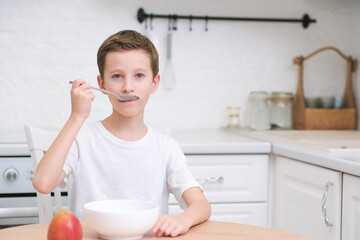 Little boy eating cereal with milk, apple for breakfast at wooden table in light white kitchen.