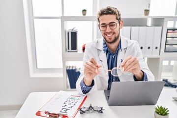 Young man optician holding glasses using laptop at clinic