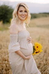 Fototapeta na wymiar Portrait of a happy pregnant blonde woman with a straw basket and a bouquet of sunflowers spending time outdoors in a wheat field at sunset. The concept of motherhood.