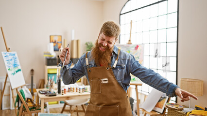 Upbeat, young redhead artist guy, joyfully swinging his paintbrush to the music in art studio,...