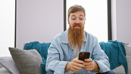Focused young redhead man, chilling on his sofa indoors, engrossed in texting on his smartphone at...