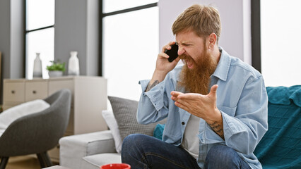 Unhappy young redhead man, argument escalating while using phone on sofa, expression shows disagreement, home setting amplifying problem, adds serious tone to indoor conversation - obrazy, fototapety, plakaty