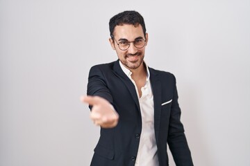 Handsome business hispanic man standing over white background smiling cheerful offering palm hand giving assistance and acceptance.
