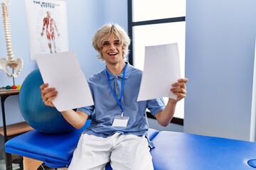 Young blond man pysiotherapist reading document sitting on massage board at rehab clinic