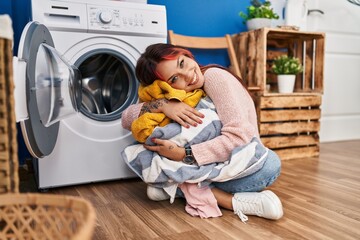 Young caucasian woman hugging clothes sitting on floor at laundry room