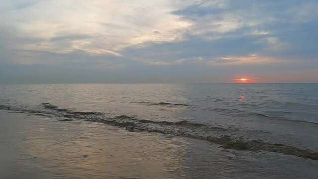This footage offers a tranquil seascape as the day ebbs into twilight. The sun, a fiery orb, dips towards the horizon of a calm sea, its fading light casting a soft glow across the water and the wet