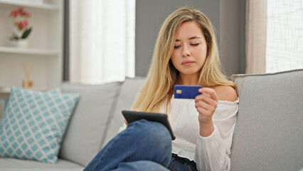 Young blonde woman shopping with touchpad and credit card sitting on sofa at home