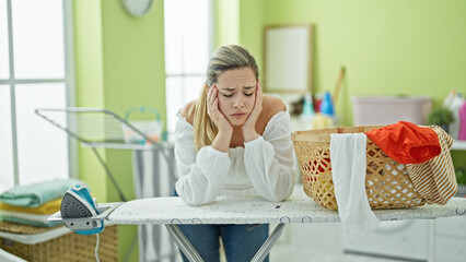 Young blonde woman leaning on ironing table with sad expression at laundry room