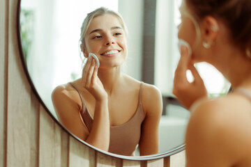 Positive attractive woman removing makeup with cotton pad, looking in mirror standing in bathroom