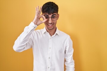 Young hispanic man standing over yellow background doing ok gesture with hand smiling, eye looking through fingers with happy face.