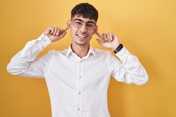 Young hispanic man standing over yellow background smiling pulling ears with fingers, funny...