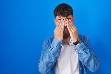 Young hispanic man standing over blue background rubbing eyes for fatigue and headache, sleepy and tired expression. vision problem