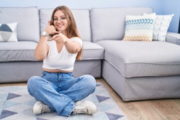 Young caucasian woman sitting on the floor at the living room smiling doing talking on the telephone gesture and pointing to you. call me.