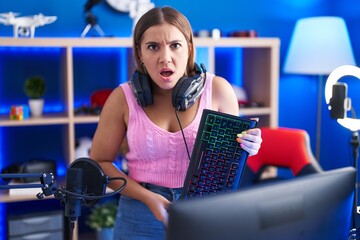 Young blonde woman playing video games holding keyboard in shock face, looking skeptical and...
