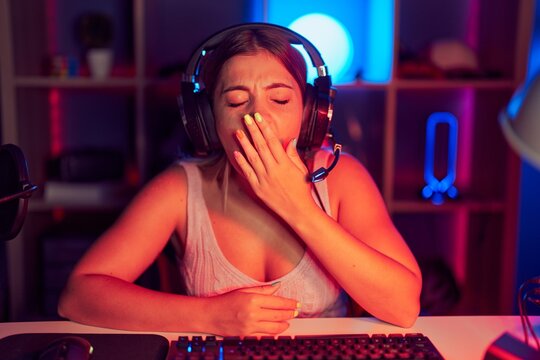 Young blonde woman playing video games wearing headphones bored yawning tired covering mouth with hand. restless and sleepiness.