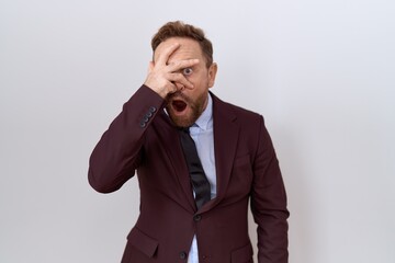 Middle age business man with beard wearing suit and tie peeking in shock covering face and eyes with hand, looking through fingers with embarrassed expression.