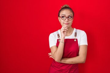 Young hispanic woman wearing waitress apron over red background with hand on chin thinking about...