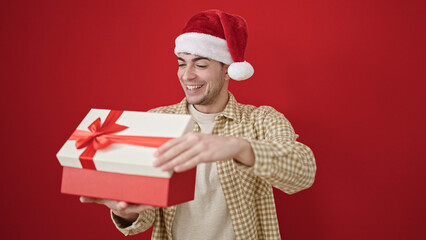 Young hispanic man wearing christmas hat unpacking gift over isolated red background