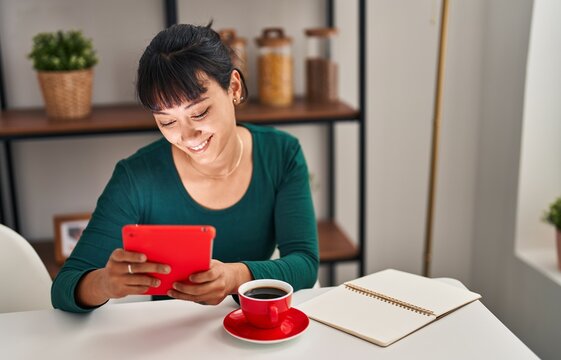 Young beautiful hispanic woman using touchpad drinking coffee sitting on table at home