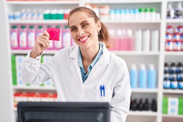 Young beautiful hispanic woman pharmacist smiling confident holding condom at pharmacy