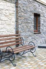 A bench with wrought-iron railings against the background of a textured stone wall of a house in the daytime.