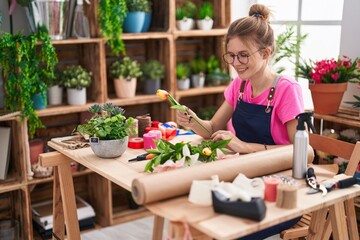 Young blonde woman florist make bouquet of flowers sitting on table at flower shop