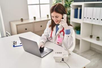 Young blonde woman doctor using laptop talking on telephone at clinic