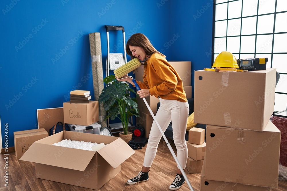 Sticker Young blonde woman singing song using paint roller as a microphone at new home