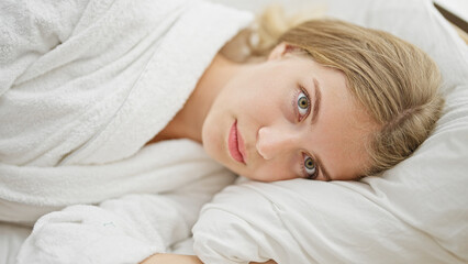 Fototapeta na wymiar Young blonde woman wearing bathrobe hugging pillow with serious face at bedroom