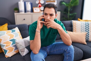 Young hispanic man talking on smartphone with worried expression at home