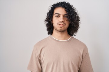 Hispanic man with curly hair standing over white background puffing cheeks with funny face. mouth...