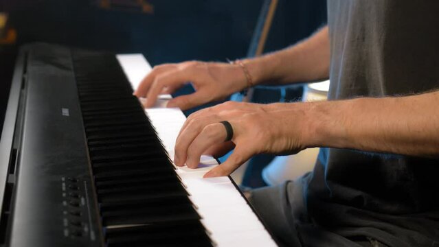The young musician's hands play the piano in a dark studio with a light at the back.