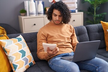 Young latin man using laptop reading notebook at home