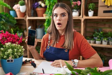 Fototapeta premium Hispanic young woman working at florist shop showing smartphone screen scared and amazed with open mouth for surprise, disbelief face