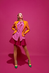 Full-length portrait of young attractive girl in unusual, strange, variegated colored outfit and posing against magenta color studio background.