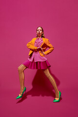 Full-length portrait of young attractive girl in unusual, strange, retro colored outfit and posing against magenta color studio background.